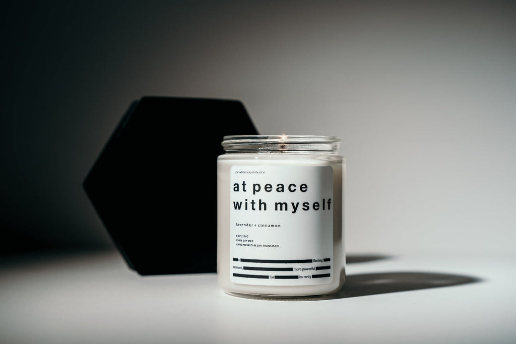 A postmodernform 'at peace with myself' candle, which is white in a clear glass jar with a white label and black text containing the candle name, scent, and a themed poem, appears in front of artistic black shapes.