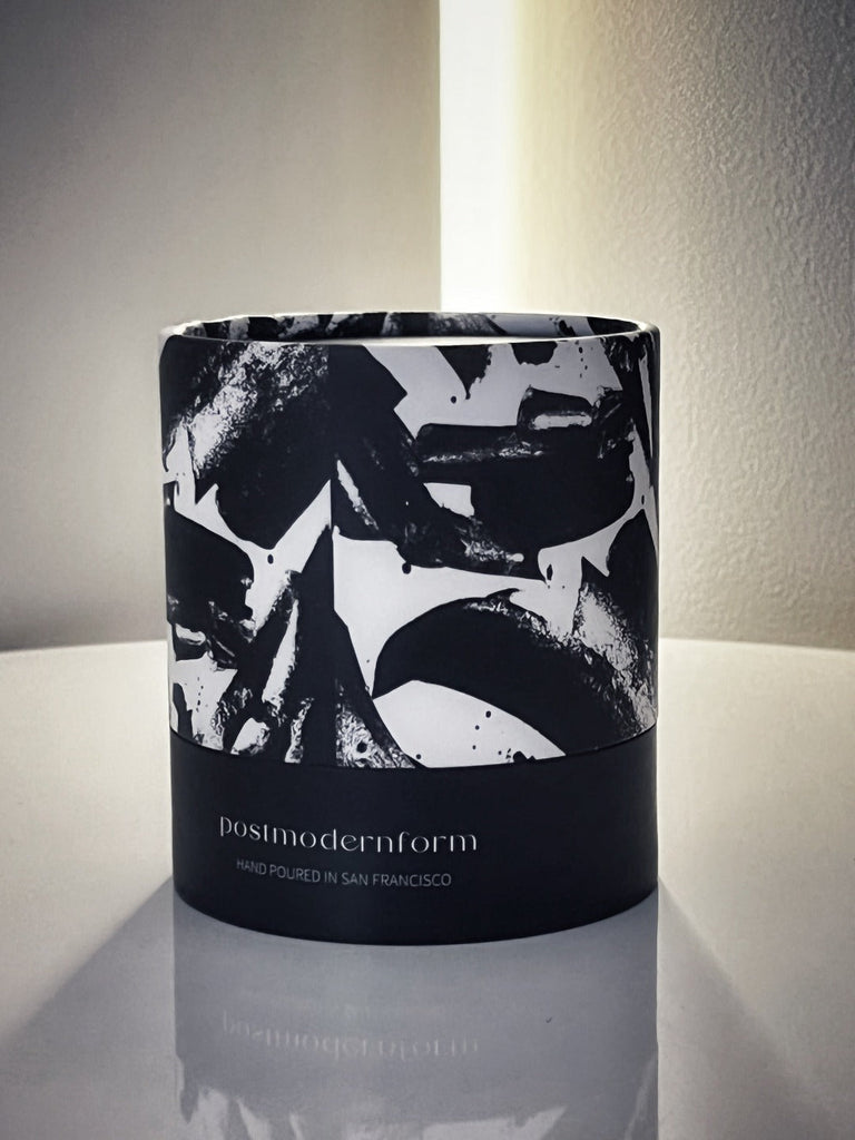 A photo of the artsy and iconic round tube packaging within which all postmodernform candles are packaged. The bottom half of the tube is black with the words 'postmodernform' and 'hand poured in san francisco' written. The top half is white with large artistic black paint strokes.