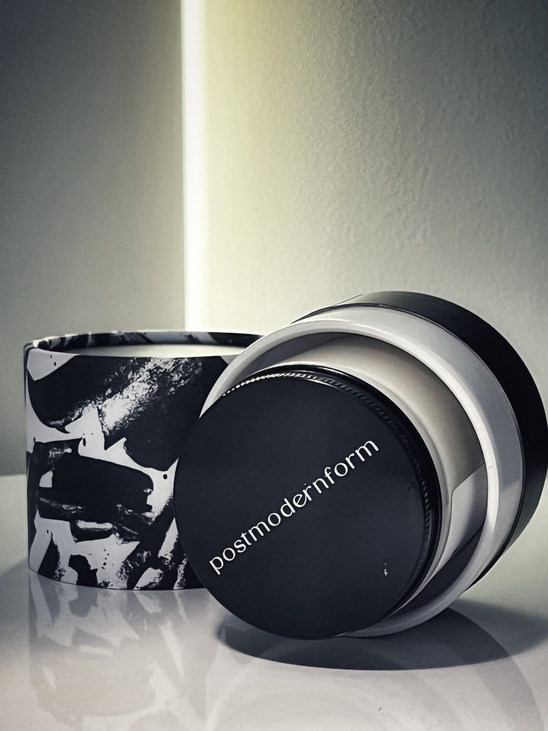 A postmodernform candle in its artsy black and white tube packaging laid on its side with the top of the candle jar shown; the brand name 'postmodernform' is displayed on the top of the candle jar.