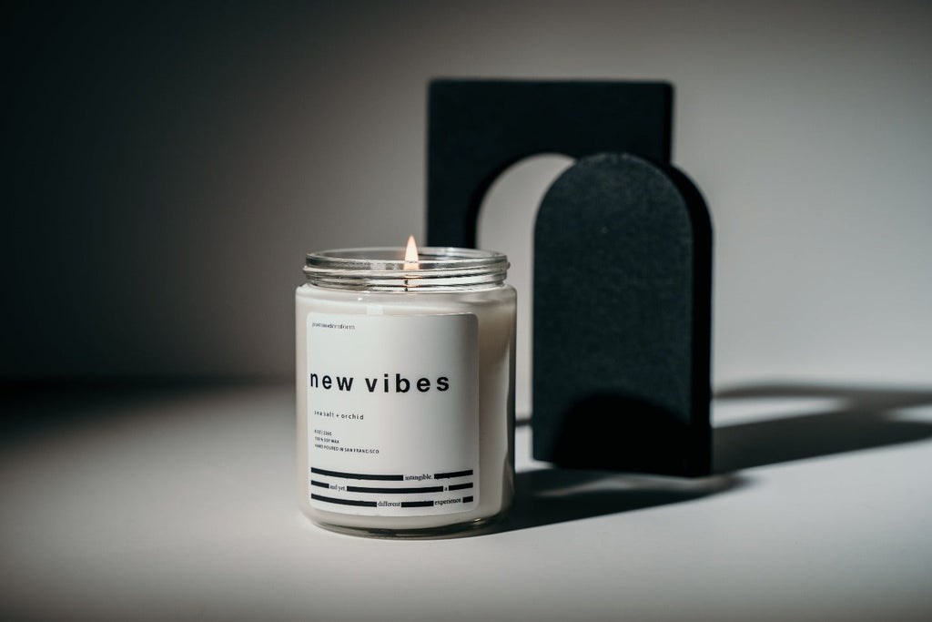 A postmodernform 'new vibes' candle, which is white in a clear glass jar with a white label and black text containing the candle name, scent, and a themed poem, appears in front of artistic black shapes.