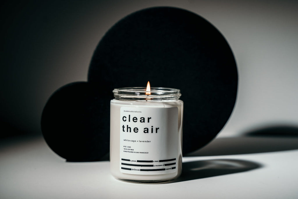 A postmodernform 'clear the air' candle, which is white in a clear glass jar with a white label and black text containing the candle name, scent, and a themed poem, appears in front of artistic black shapes.